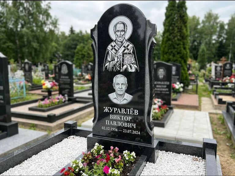 Памятник с гравировкой иконы: стела с образом в полный рост на обороте — гравировка образа Матроны Московской, низкие кусты спиреи в цвету, работа «Лебедева» в Оренбурге